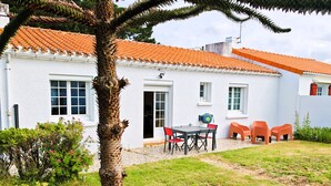 A white building with a tiled roof, a door, and windows. There is a table with chairs and a small garden area with grass and plants. - Les Chouans - Holiday home (Les Sables-d´Olonne)