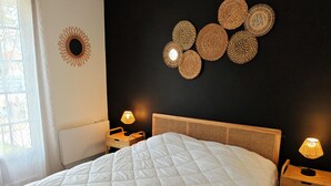 A neatly made bed with a white quilt, a dark wall with woven wall art, and two bedside lamps. - Les Chouans - Holiday home (Les Sables-d´Olonne)