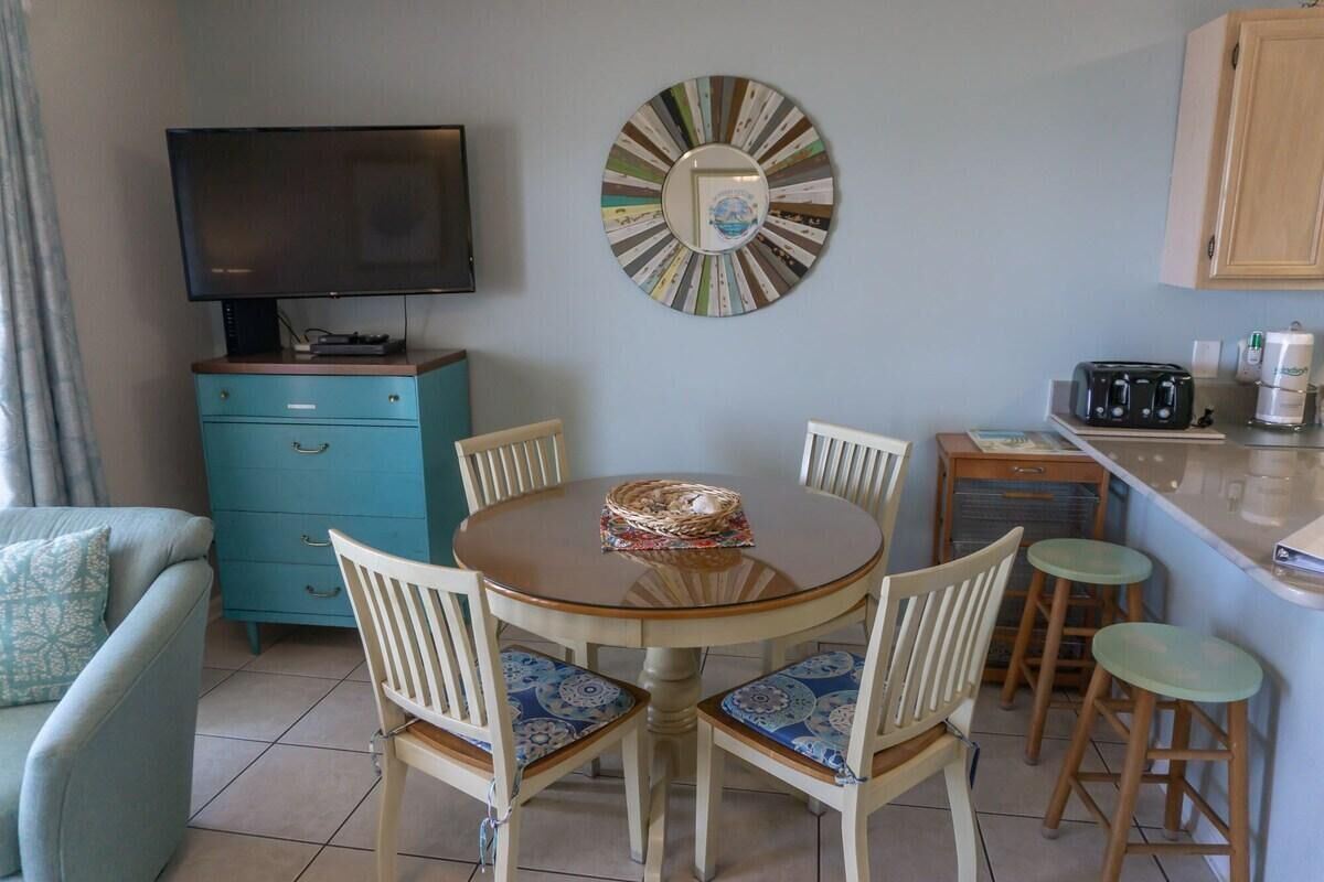 A dining area with a round table, chairs, a television, a decorative mirror, and a kitchenette.