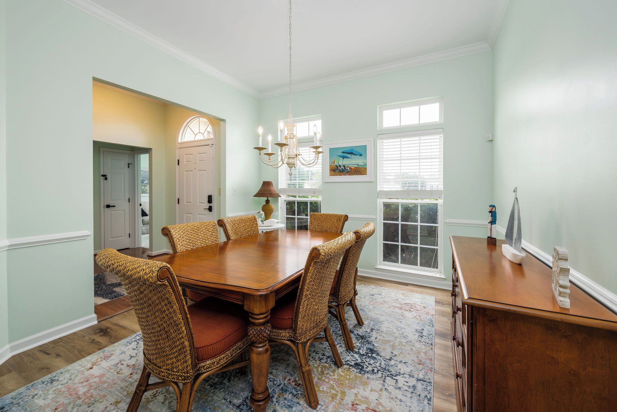 A dining room with a wooden table and wicker chairs, a chandelier, and a window with blinds.