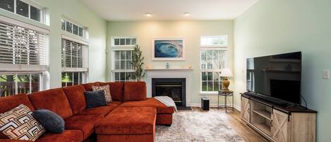 A living room with a sectional sofa, a fireplace, a television, and a large window with blinds.