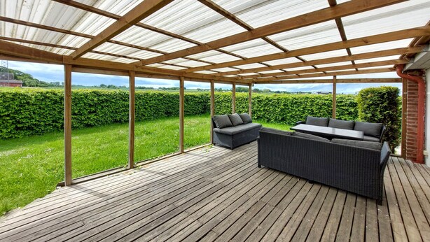 A covered outdoor patio with wooden flooring, a seating area with a sofa and chairs, and a view of a green landscape. - 2 bedroom beautiful home in Fleninge (Fleninge)