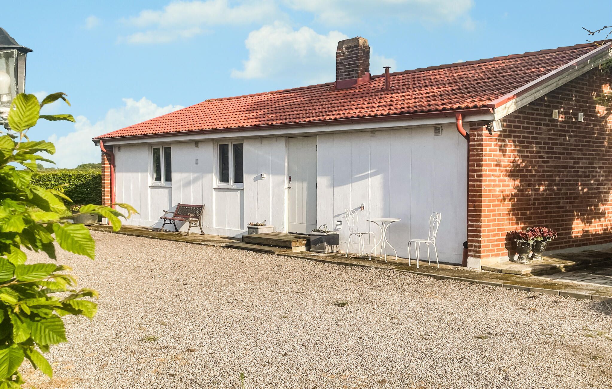 A single-story house with a red-tiled roof, white exterior walls, and a gravel driveway. There are outdoor seating arrangements and a small garden area visible.