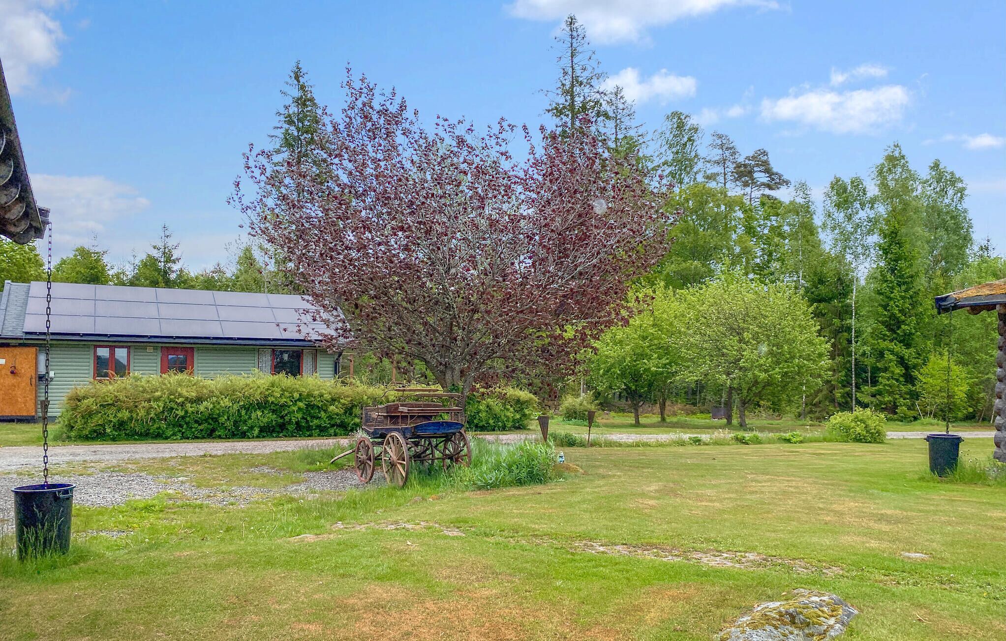 Une cabane rustique à l’extérieur vert et au toit en pente, entourée d’une pelouse bien entretenue et de divers arbres, dont un arbre à fleurs particulièrement remarquable.