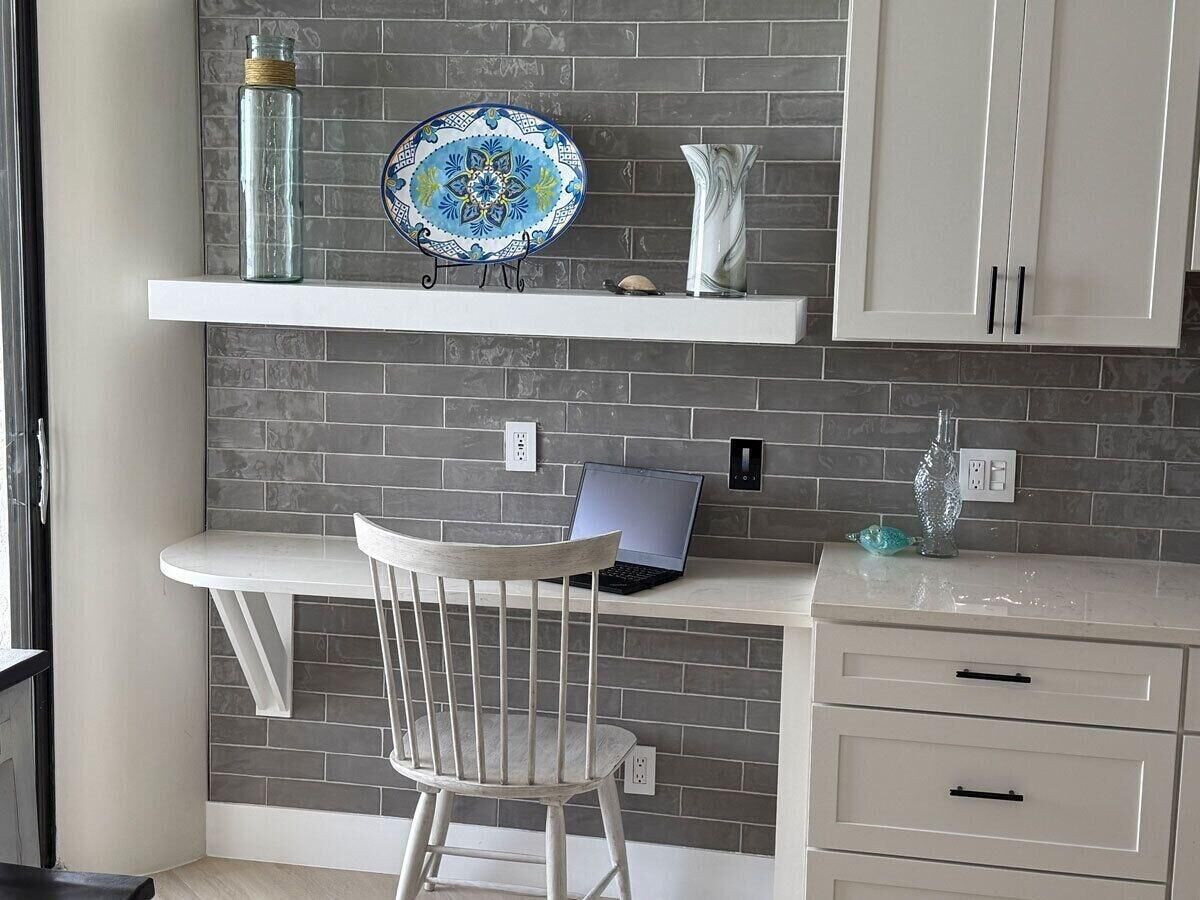 Une cuisine avec un ordinateur portable posé sur un plan de travail blanc, une chaise blanche, une assiette décorative et un vase.
