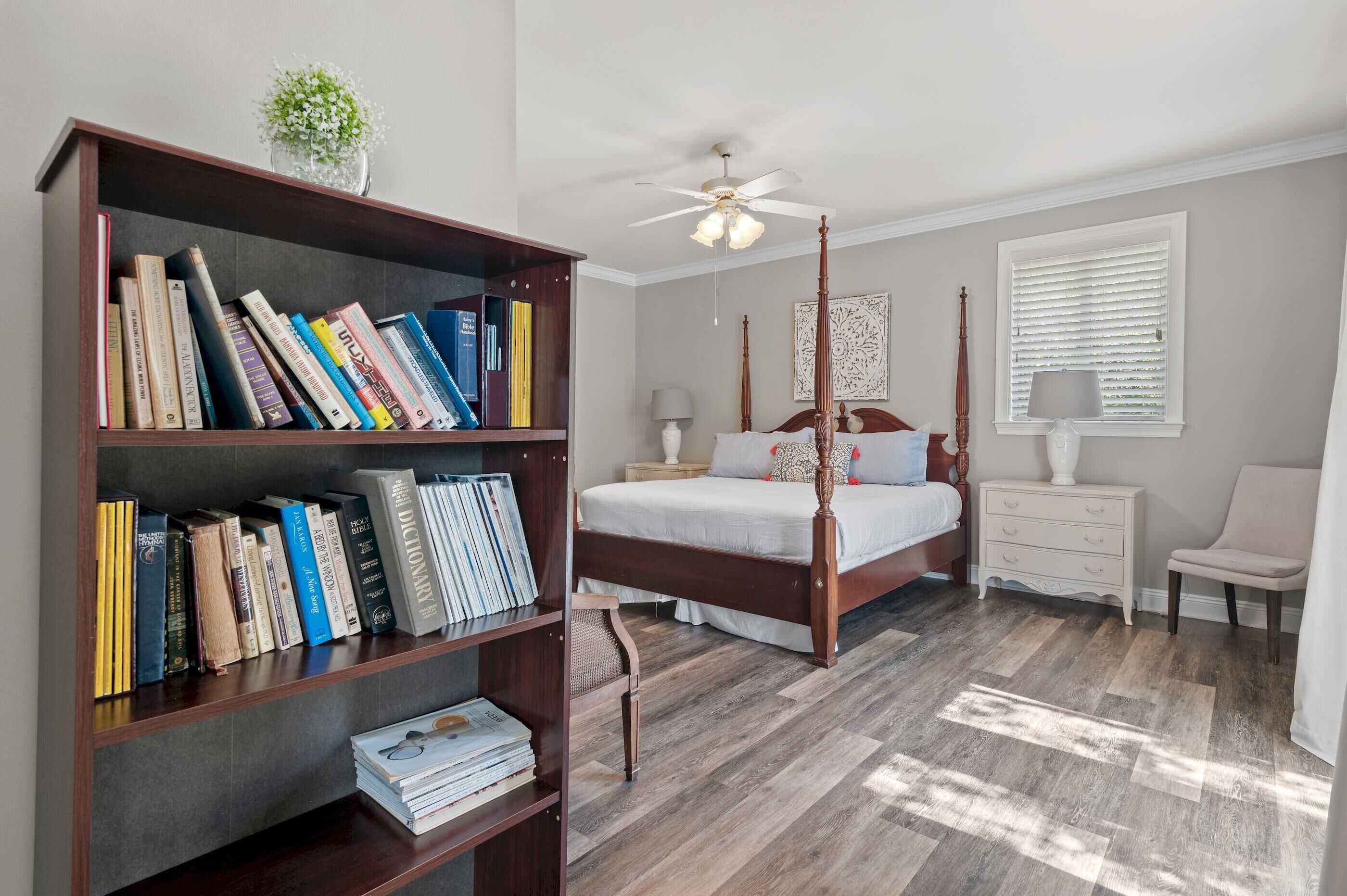 Ein Schlafzimmer mit Himmelbett, Bücherregal, Nachttisch und Fenster mit Jalousien.