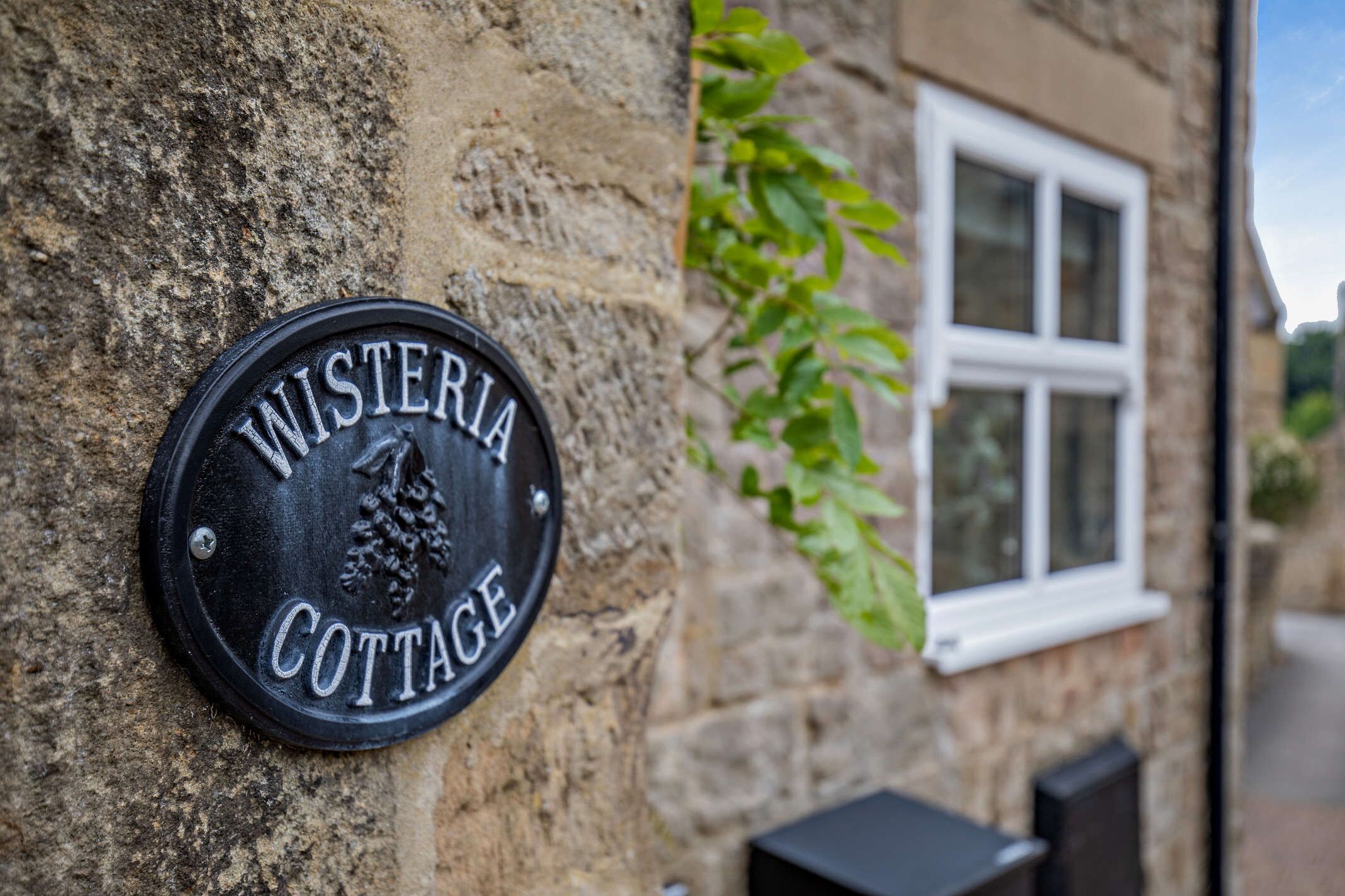 A black metal plaque on a stone wall reading "WISTERIA COTTAGE."