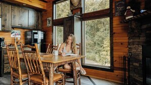 A rustic cabin interior with wooden walls, a large window overlooking a forest, a dining table with chairs, and a kitchen area with appliances. - Elk Ridge Cabin – Private Hot Tub & Stunning Views (Bryant)