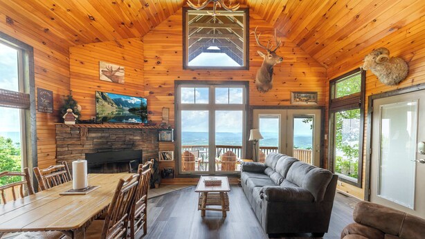 A log cabin interior with a dining area, fireplace, and a large window overlooking a scenic view. - Elk Ridge Cabin – Private Hot Tub & Stunning Views (Bryant)