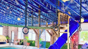 An indoor pool area with a blue slide, rope climbing net, and a stone wall in the background. - Elk Ridge Cabin – Private Hot Tub & Stunning Views (Bryant)