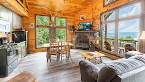 A wooden cabin interior with a dining area, fireplace, and large windows overlooking a scenic view. - Elk Ridge Cabin – Private Hot Tub & Stunning Views (Bryant)