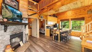L’intérieur d’un chalet comprend une cheminée en pierre, un coin repas en bois, une cuisine et un balcon offrant une vue sur la forêt.
