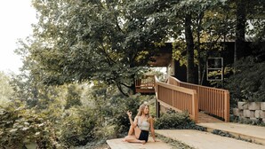 Eine Person sitzt auf einer Holzdiele in einem Garten mit einer Holztreppe und einem Haus im Hintergrund.