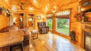 Ein gemütliches Blockhäuschen-Interieur mit Holzwänden, einem Kamin, einer komfortablen Sitzecke und einem großen Fenster mit Blick ins Freie.