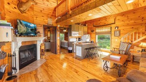 Una cabaña amplia con chimenea, pisos de madera, cocina, comedor y un altillo con balcón.