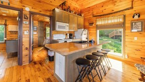 Interior de una cabaña de madera con una zona de cocina que incluye un microondas, estufa y fregadero. Hay tres taburetes de bar cerca de la isla de cocina.