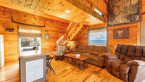 Interior de una cabaña de madera con una escalera, un sofá, una mesa de comedor y un refrigerador.