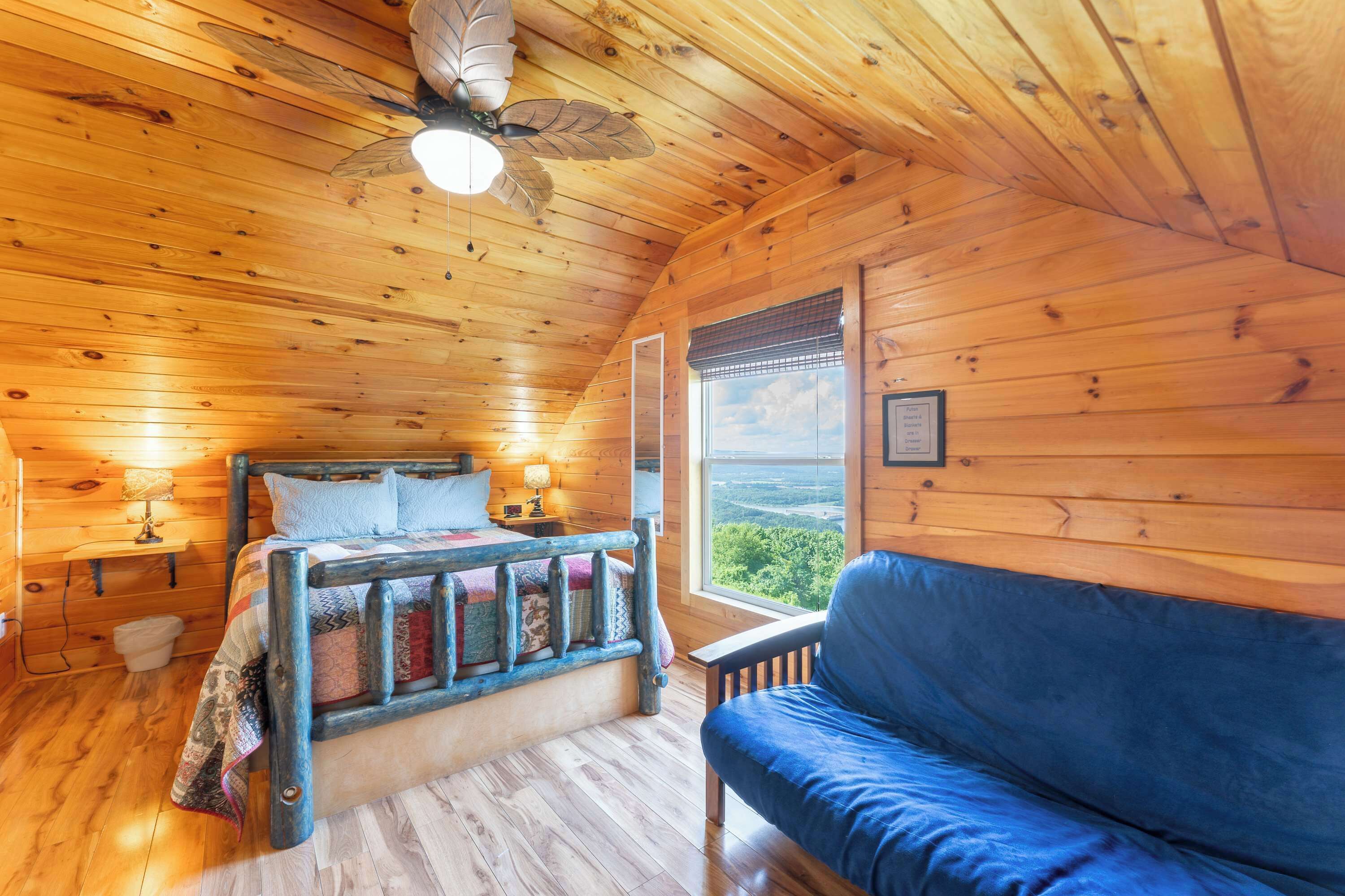 A wooden cabin bedroom with a bed, a blue couch, a ceiling fan, and a window with a view.