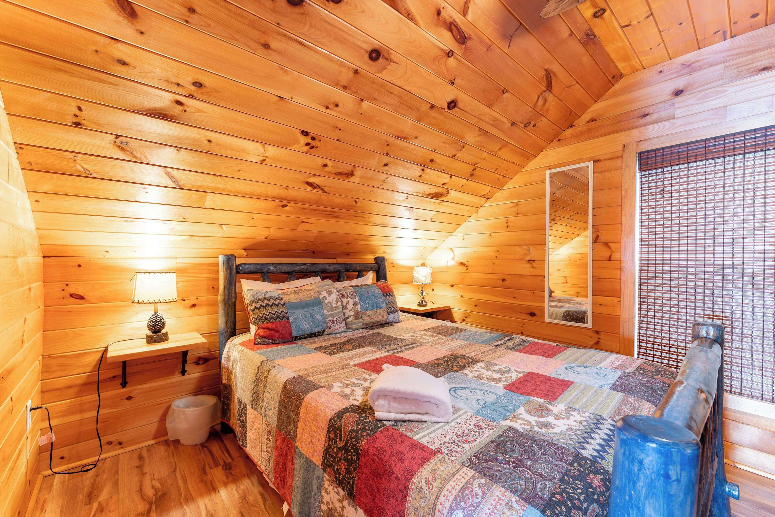 A cozy wooden cabin bedroom with a bed, two bedside lamps, a wooden headboard, a window with blinds, and a small wooden stool.