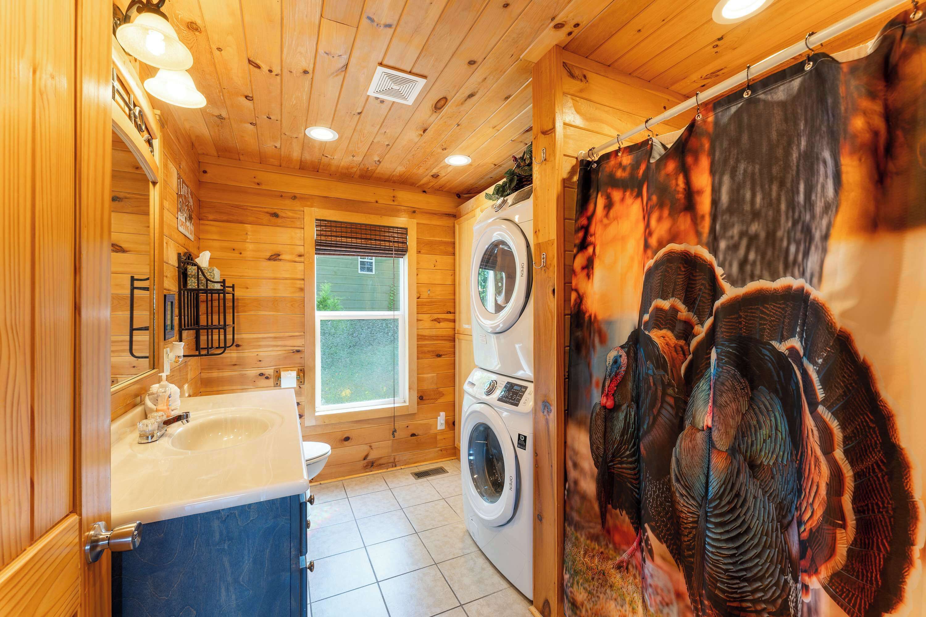 A wooden cabin bathroom with a sink, two stacked washing machines, and a shower curtain with a bird design.