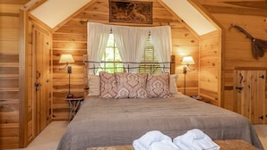 A cozy wooden room with a bed, two bedside lamps, a painting, and a wooden bench with towels. - The Cabins at Seven Foxes (Lake Toxaway)