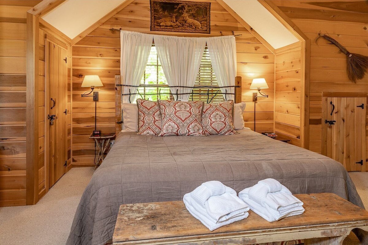 A cozy wooden room with a bed, two bedside lamps, a painting, and a wooden bench with towels.