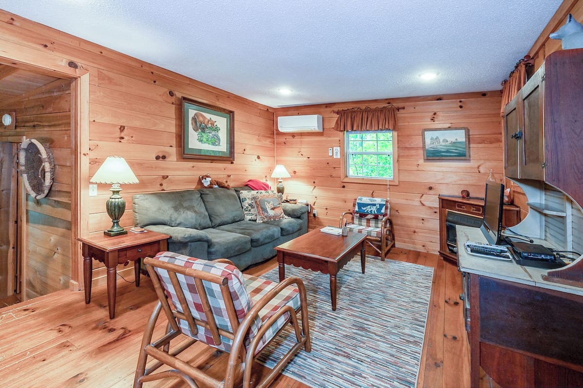 A cozy cabin interior with wooden walls, a sofa, a rocking chair, a coffee table, and a small kitchen area with a refrigerator and a microwave.
