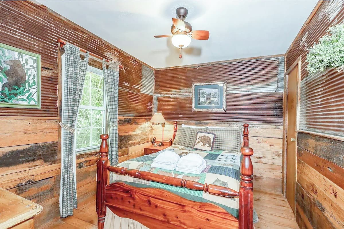 A rustic wooden room with a four-poster bed, a ceiling fan, and a window with curtains.