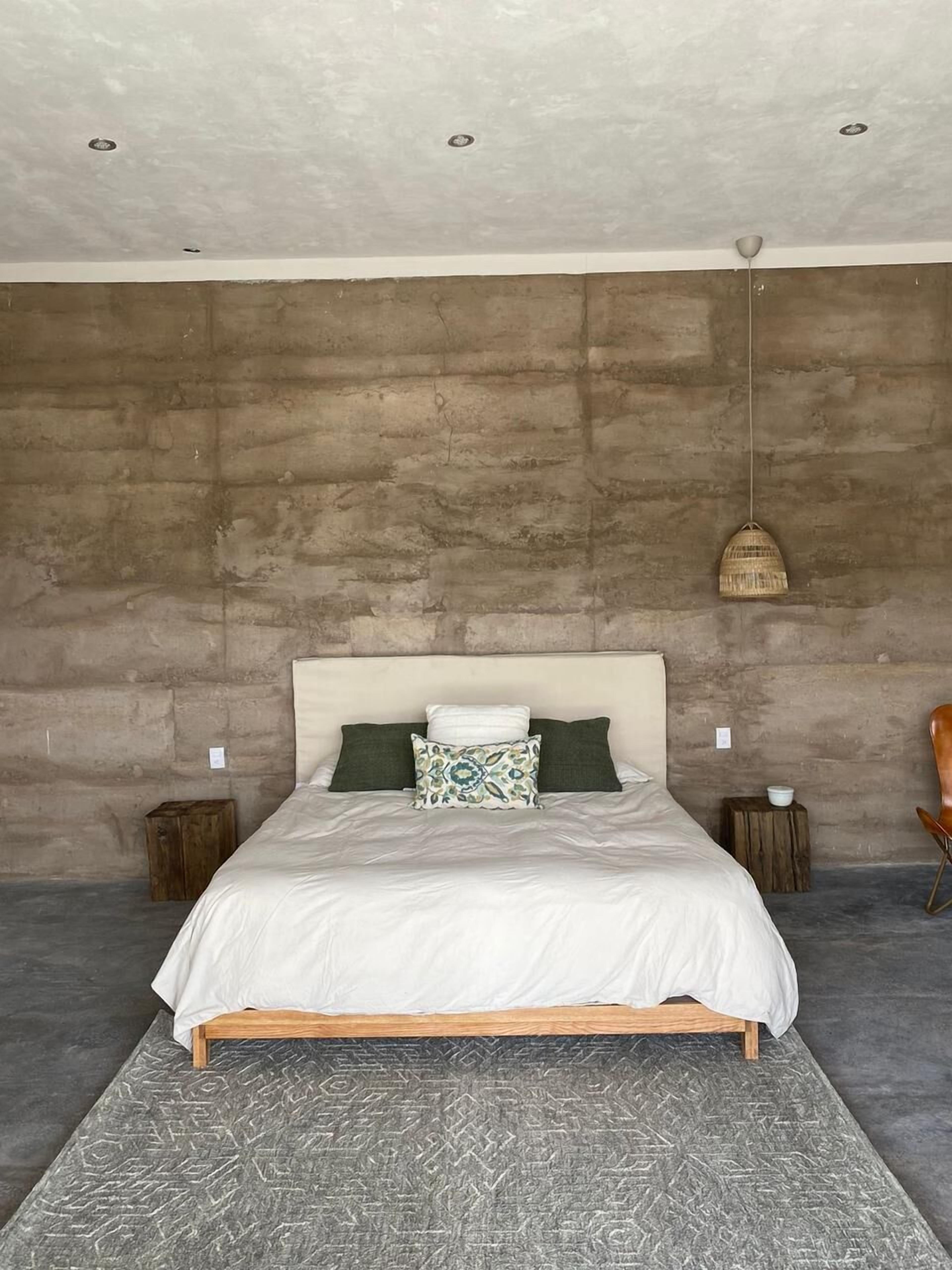 A bedroom with a concrete wall, a wooden bed frame, a white bedspread, and a patterned rug.