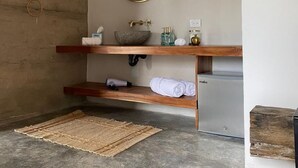 Un baño con pared de madera, un espejo redondo, una repisa de madera con artículos de aseo y una alfombra tejida en el suelo.