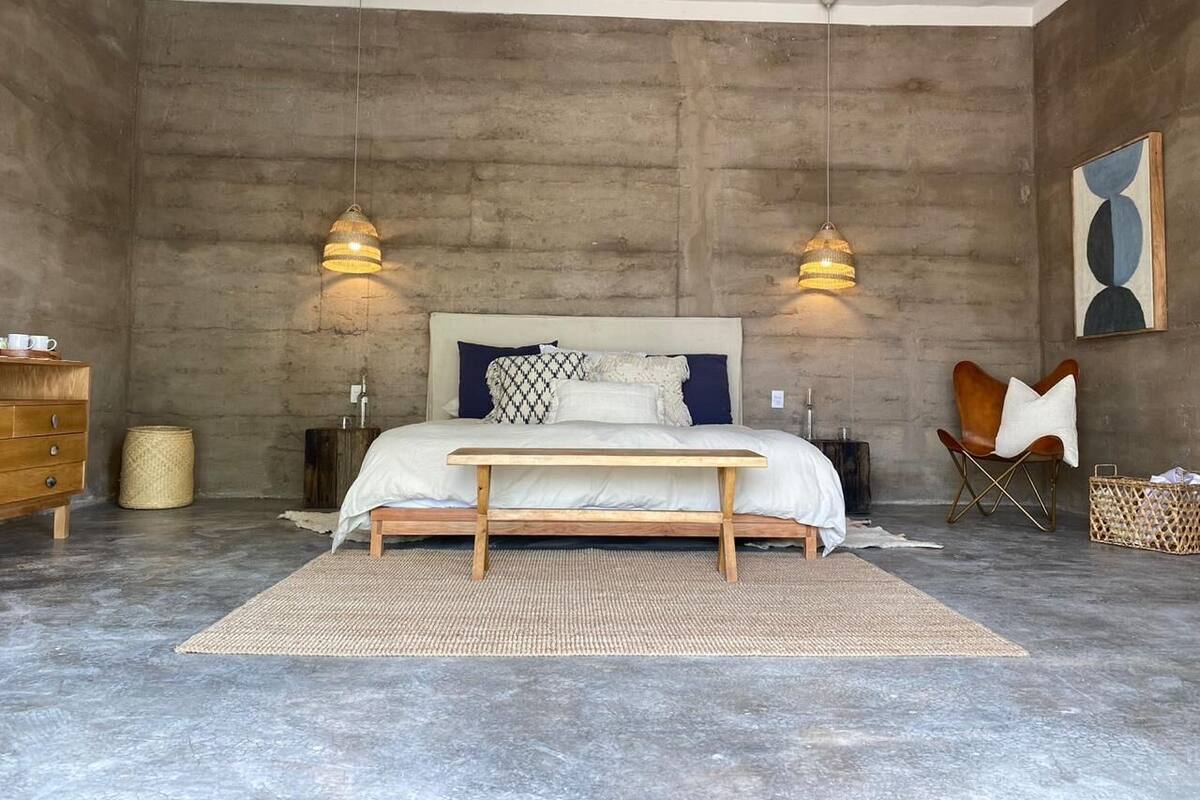 A bedroom with a large bed, two bedside lamps, a wooden bench, a woven rug, and a leather chair.