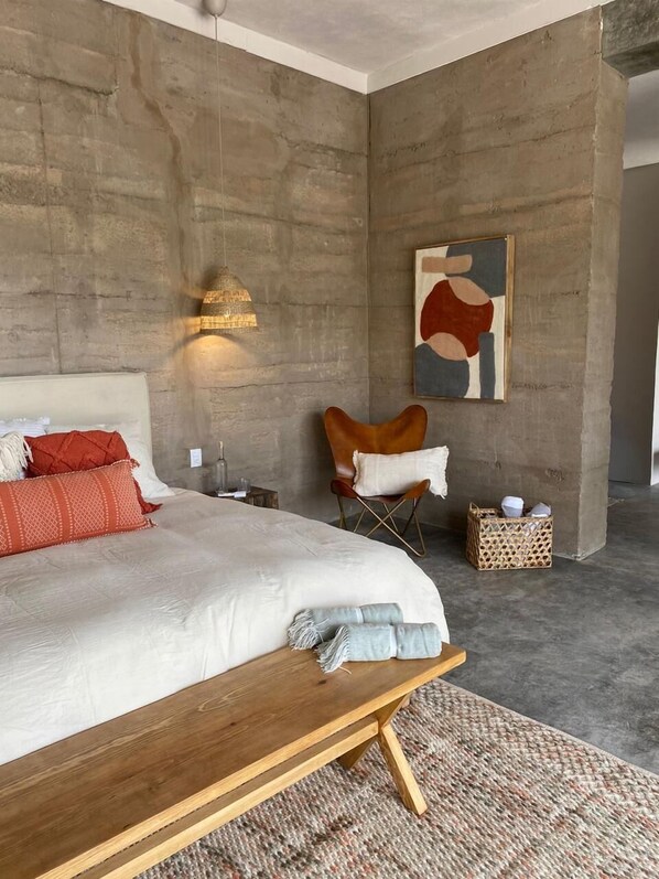 Un dormitorio moderno con una cama grande, un banco de madera, una silla, una lámpara de pared y un cuadro.