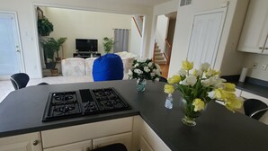 Eine Küche mit Herd, einem Esstisch und einer Vase mit Blumen. Eine Treppe führt zu einem Wohnbereich mit einer Couch und einem Fernseher.