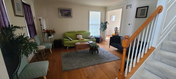 A living room with a green sofa, a black sofa, a wooden coffee table, and a grey rug. There is a staircase with a wooden handrail leading to a landing.