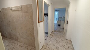A hallway with beige tiled flooring, a mirror on the wall, and a door leading to a kitchen area with a refrigerator and a chair. - Vacation Apartment (GF) at Grossensee - Relaxation Close to Hamburg (großensee)