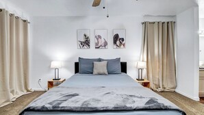 Une chambre à coucher comprenant un lit, deux tables de chevet avec des lampes, un ventilateur de plafond et trois œuvres d’art accrochées au mur.