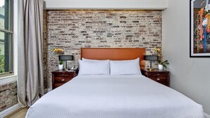 Habitación de hotel con cabecera de ladrillo, una cama grande con ropa blanca, dos mesitas de noche con lámparas y una ventana con cortinas.
