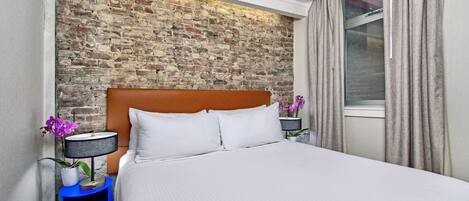Une chambre d’hôtel avec une tête de lit en briques, un lit recouvert de draps blancs, une table de chevet bleue avec une lampe et une fenêtre avec des rideaux.