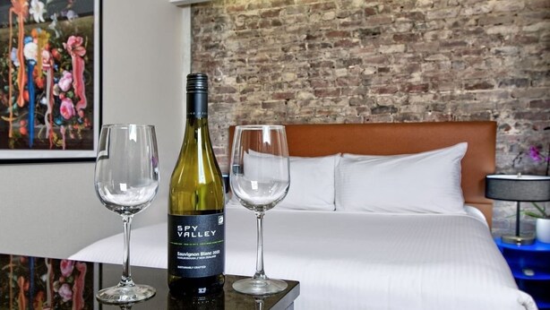 A hotel room with a bed, a brick wall, and a painting. There is a bottle of wine and two glasses on a table.