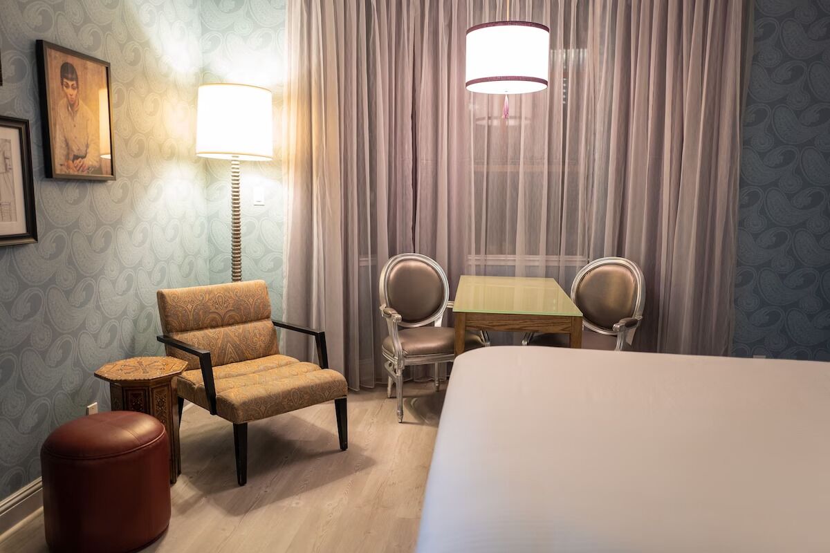 Quarto de hotel com uma cama, duas cadeiras, uma mesa, um abajur e um banquinho.