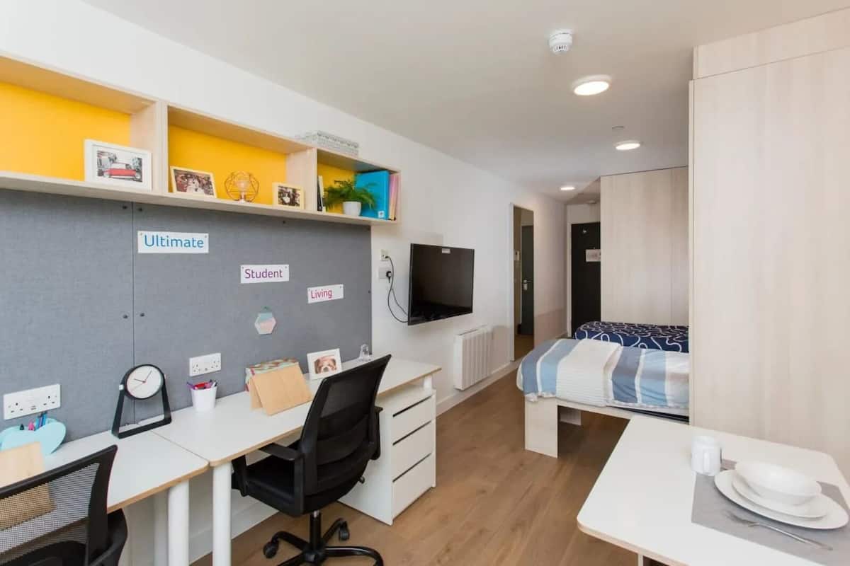 A modern, well-lit room with a desk, chair, and bed. There are shelves with personal items and a TV mounted on the wall.