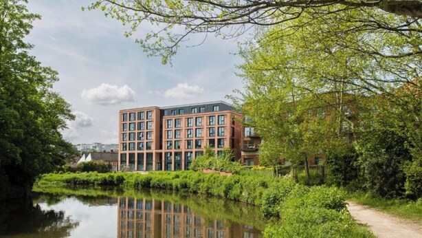 A modern building with a reflective water body in front, surrounded by greenery and a path.