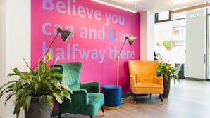 A modern waiting area with a motivational wall, green and orange chairs, and potted plants.