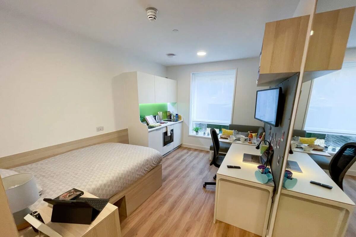 A compact room with a bed, a desk, a chair, a small kitchenette, and a TV.