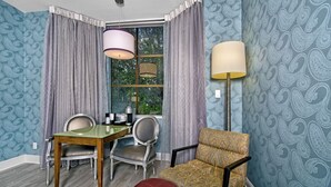 Ein Zimmer mit einem Esstisch, zwei Stühlen, einem roten Hocker, einem gemusterten Sessel und einer Stehlampe.