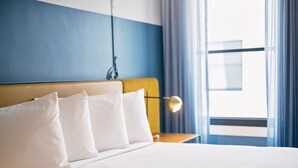 Ein Hotelzimmer mit Bett, weißen Kissen, einem Kopfteil, einem Nachttisch mit Lampe und einem Fenster mit Vorhängen.