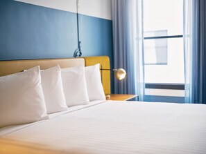 Habitación de hotel con cama, almohadas blancas, cabecera, mesita de noche con lámpara y ventana con cortinas.