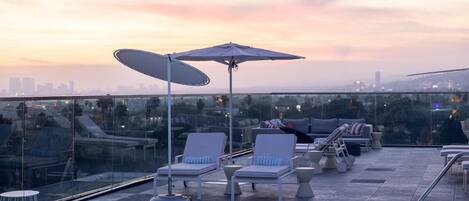 Zona de piscina en la azotea con sillones de descanso y sombrillas, con vistas a un horizonte urbano al atardecer.
