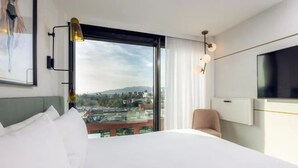 Habitación de hotel con una cama grande, un televisor montado en la pared, una silla y vista a un paisaje urbano a través de un ventanal amplio.