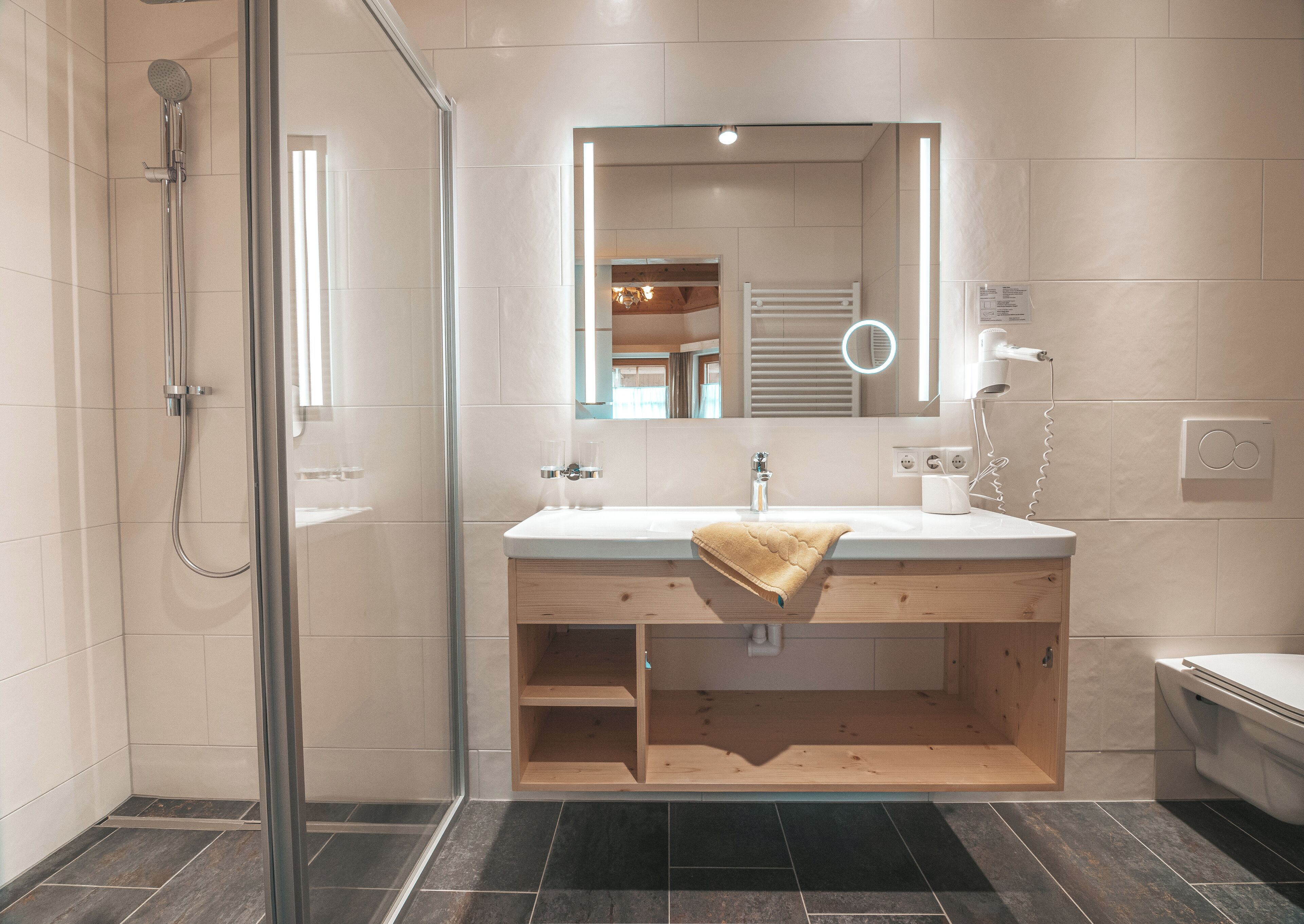 A modern bathroom with a glass shower enclosure, a wooden vanity, and a large mirror with integrated lighting.